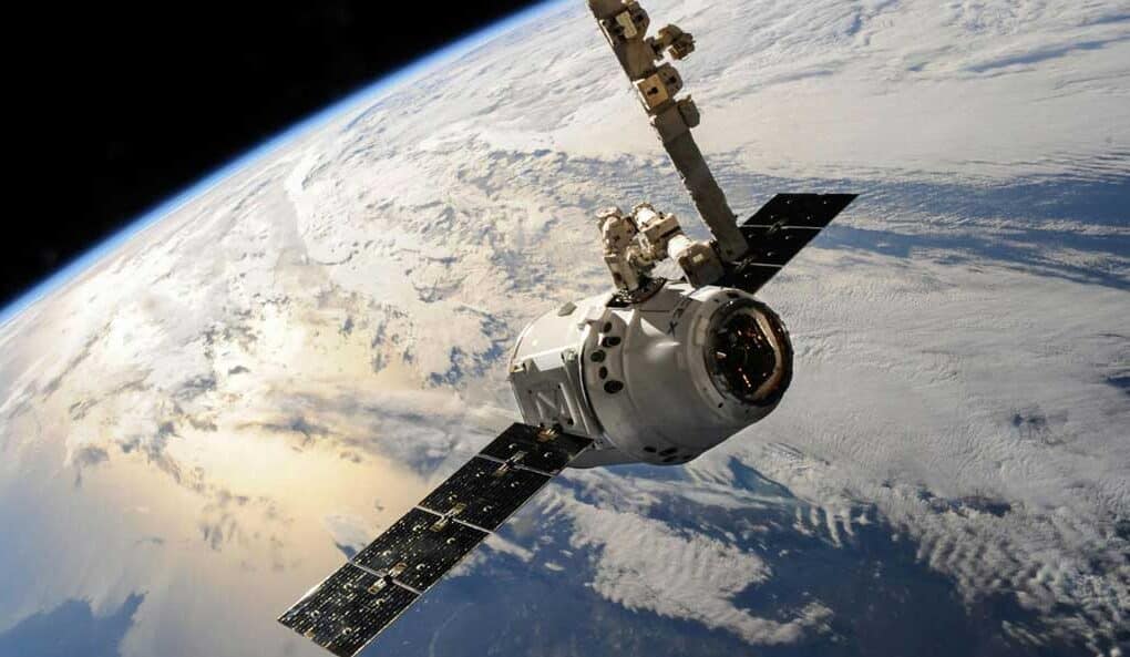 A satellite or spacecraft, supported by an aerospace logistics partner, orbits above Earth with solar panels extended and the planet’s cloud-covered surface visible in the background.